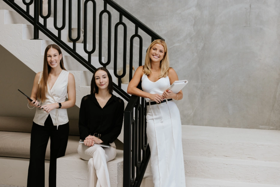 Three female marketing professionals smiling at camera next to stairway